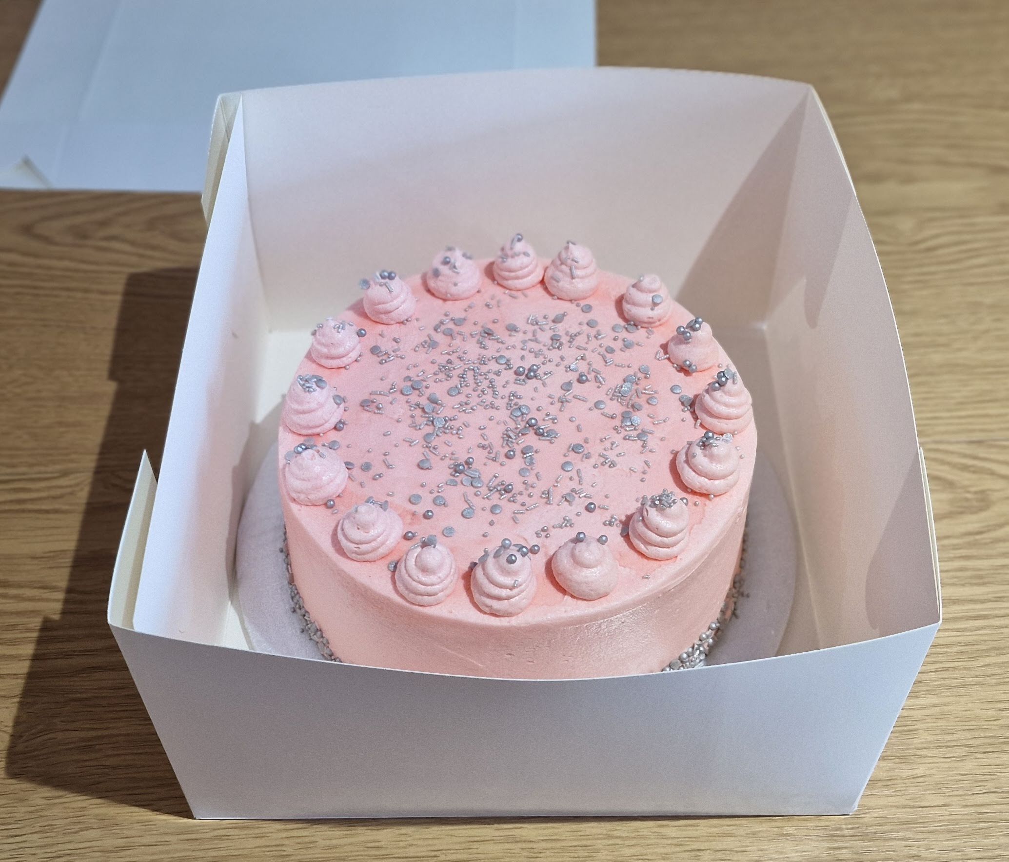 Bespoke round cake featuring pink strawberry icing swirls and silver sprinkles inside a cake box, handcrafted by Fontaines Fancies in Greenford