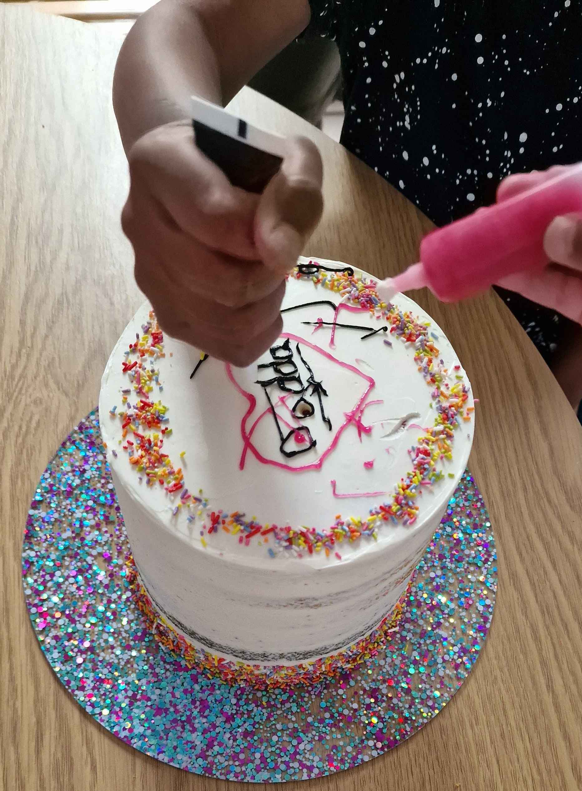 Hands-on process of adding pink and black icing writing and sprinkles to a fresh custom cake in our Fontaines Fancies Greenford studio kitchen